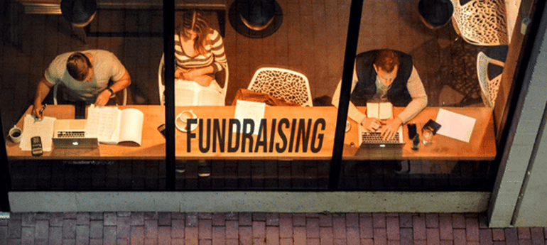 customers sit at a bar reading and looking at laptops in a cafe with the word fundraising on the window